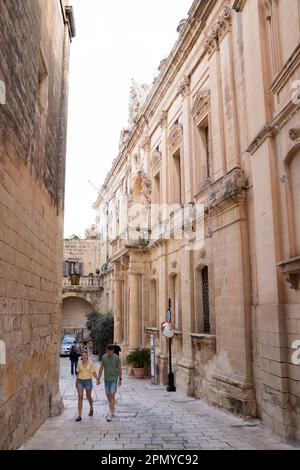 Mdina, Malta - November 13, 2022: Limestone buildings in the paved streets of the quiet city and a tourist couple walking sightseeing Stock Photo