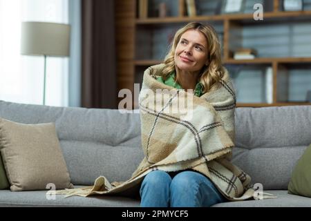 An elderly woman covered with a blanket sits dreamily at home on the sofa and rests. Stock Photo
