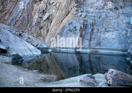 Reflections in the pool beneath a dry waterfall surrounded by rocks with sloping rock on the left. Stock Photo