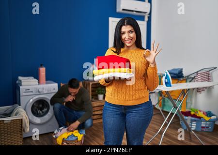Young couple doing laundry at home doing ok sign with fingers, smiling friendly gesturing excellent symbol Stock Photo