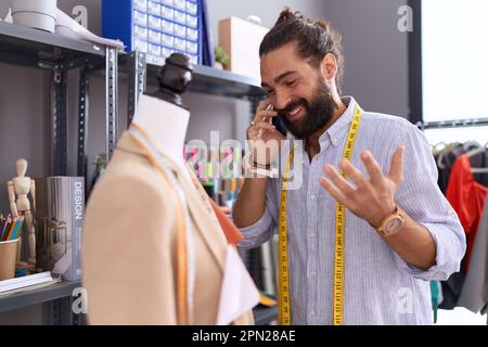 Young hispanic man tailor talking on smartphone standing by manikin at atelier Stock Photo