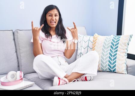 Young hispanic woman sitting on the sofa at home shouting with crazy expression doing rock symbol with hands up. music star. heavy concept. Stock Photo