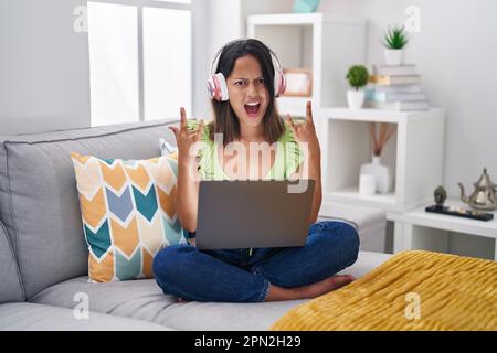 Hispanic young woman using laptop at home shouting with crazy expression doing rock symbol with hands up. music star. heavy concept. Stock Photo