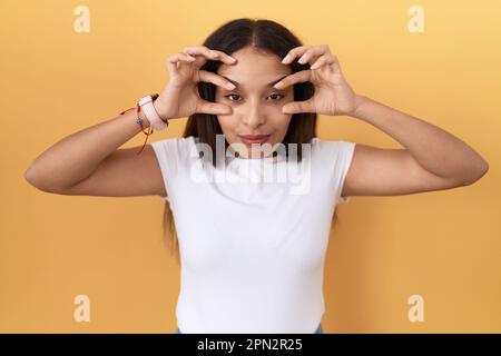 Young arab woman wearing casual white t shirt over yellow background trying to open eyes with fingers, sleepy and tired for morning fatigue Stock Photo