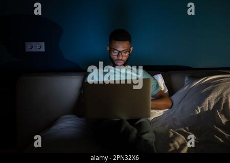 African American guy freelance lying in bed using laptop working remotely until late hours Stock Photo