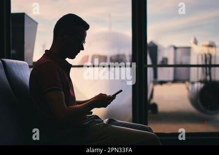 Traveler using phone while waiting for flight. Silhouette of man waiting by window of airport terminal behind which is airplane. Stock Photo