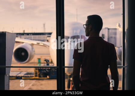 Traveler is looking out of airport window at airplane. Silhouette of man waiting for his flight. Stock Photo