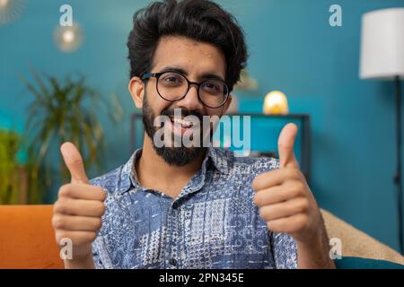 Like. Happy excited indian man looking approvingly at camera showing thumbs up, like sign positive something, good great news, positive feedback. Young hindu guy sitting on couch at home living room Stock Photo