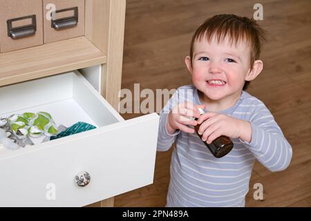Toddler baby opened a cabinet drawer with pills and a vial of potion. Child boy holding medicine bottle standing in home living room. Kid age one year Stock Photo