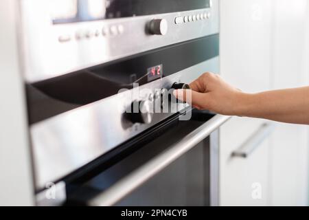 Unrecognizable Woman Using Electic Oven In Kitchen, Adjusting Temperature With Hand Stock Photo