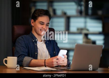 Cheerful young man entrepreneur sitting at workdesk, using laptop, smartphone Stock Photo