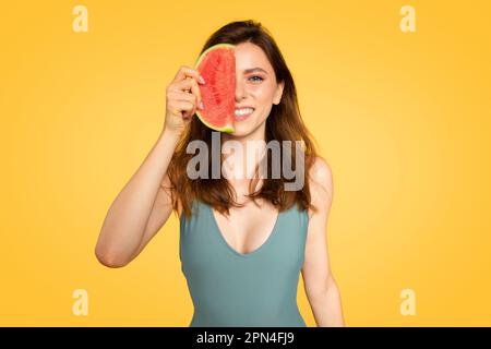 Summer time concept. Pretty woman holding half of red watermelon near face, posing over yellow background Stock Photo