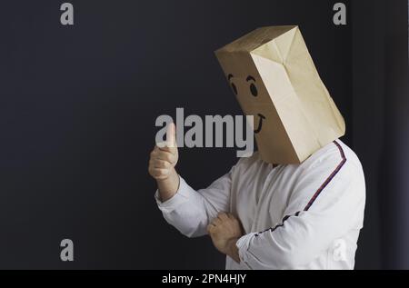 A man in a white shirt with a paper bag on his head, with a smiley face drawn, stands against the wall, overwhelmed with feelings, shows a hand gestur Stock Photo
