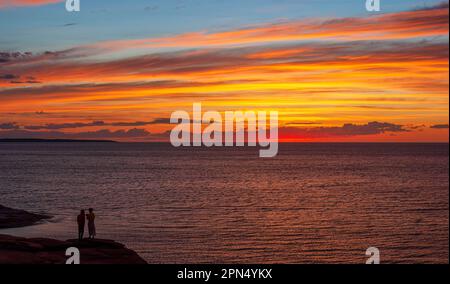 Young couple enjoying a colorful sunset on the Gulf of St Lawrence coast. Dramatic sky in red and orange shades. Oceanview Lookoff, PEI National Park. Stock Photo