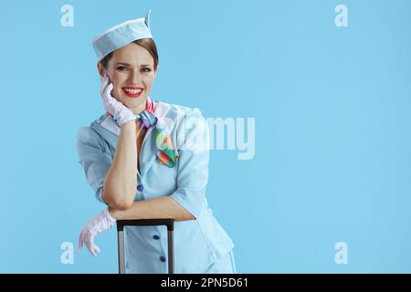 happy elegant female air hostess against blue background in blue uniform with trolley bag. Stock Photo
