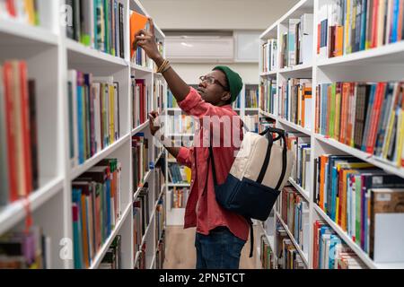 Nerd student african american man choosing book in university library taking it from shelf. Stock Photo