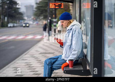 Stylish guy waiting bus sitting on bus stop bench in downtown with coffee browsing phone. Stock Photo