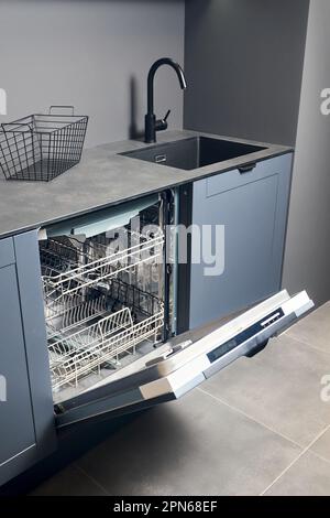 Opened empty dishwasher machine in contemporary blue grey kitchen with compact high pressure laminate HPL countertop and black square sink and tiled Stock Photo