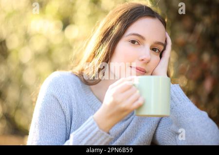 Pensive woman sitting in a park holding coffee looks at you at sunset Stock Photo