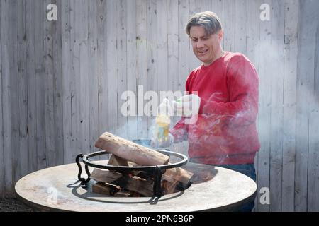 Round grill in the shape of a bowl with fire inside. A man prepares a hearth for barbecue. Firewood is burning in the bowl of the outdoor hearth. Stock Photo