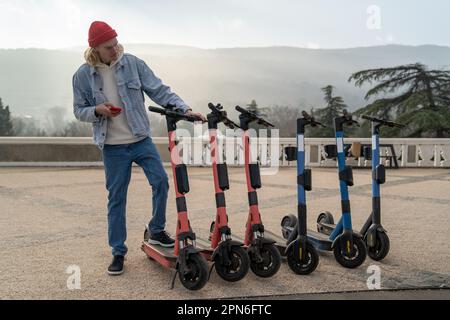 Guy hipster choosing electric scooter for rent in city park activating using smartphone. Stock Photo
