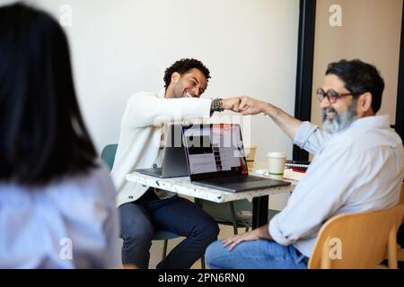 Happy male entrepreneurs giving fist bump while sitting in cafe Stock Photo
