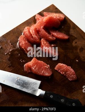 Peeled and sliced, divided in sections grapefruit on a wooden board with a knife next to it. The board rests on the marble countertop Stock Photo