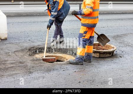 A team of road workers in orange-and-blue overalls with a shovel and brush clear a site for a new sewer manhole on the roadway. Copy space. Stock Photo