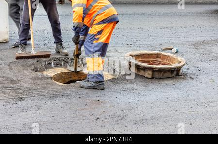 A team of road workers in orange-and-blue overalls is repairing a sewer manhole on the carriageway. Copy space. Stock Photo