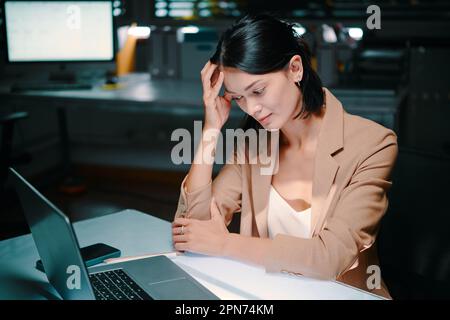 Tired businesswoman sitting at desk in dar office and pondering over problem Stock Photo