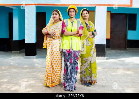 confident young indian female engineer wearing yellow hard hat and vest standing cross arms along with two other women. concept of woman empowerment. Stock Photo