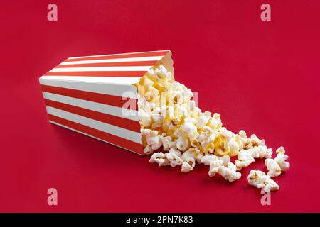Tasty cheese popcorn falling out of a red striped carton bucket, isolated on red background. Scattering of popcorn grains. Movies, cinema and entertai Stock Photo