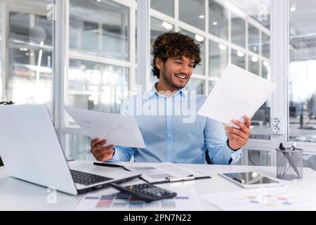 Successful financier accountant working inside office with laptop and documents, man satisfied with audit results smiling and reading financial reports, businessman using laptop for paperwork. Stock Photo