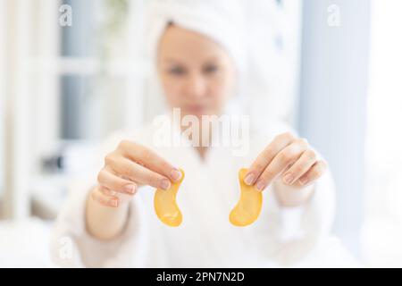 Focus on pair of golden eye patches being held by young caucasian woman wearing bathrobe and soft towel on head in home interior. Anti-aging under-eye masks fighting dark circles and fine lines. Stock Photo