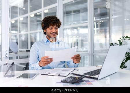 Successful financier accountant working inside office with laptop and documents, man satisfied with audit results smiling and reading financial reports, businessman using laptop for paperwork. Stock Photo