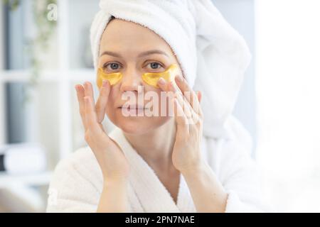 Close up view of lovely caucasian female in towel and dressing gown applying pair of eye patches after cleansing procedures in morning routine. Pretty woman taking care of delicate skin around eyes. Stock Photo