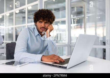 Upset young businessman working inside office with laptop, man fails to complete technical project on time, hispanic man frustrated sitting at desk. Stock Photo