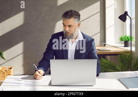 Mature businessman working in the office, using his laptop, and writing something on paper Stock Photo