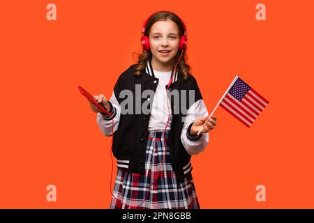 Teen school girl in casual clothes and headphones holds small American flag, or the U.S. flag and smartphone. Languages courses online. Learning Ameri Stock Photo