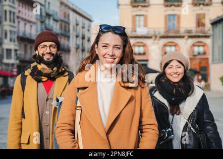Group of young people looking at camera on european city street. Portrait of three cheerful multiethnic friends standing outdoors on a journey. Front view of few happy hipsters enjoying together. High quality photo Stock Photo