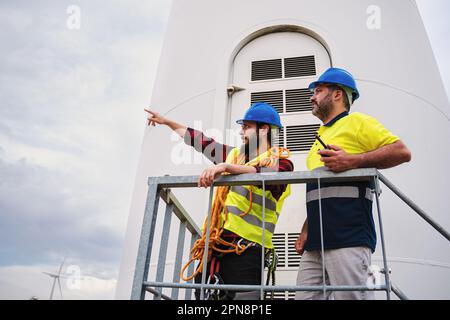 Two windmill engineer pointing to the turbine they have to do maintenance on. Renewable energy technicians expert taling, working together and doing an inspection of the generator checking the power. High quality photo Stock Photo