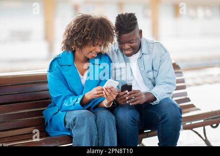 Cheerful Black Couple Using Smartphones While Sitting On Bench Outdoors Stock Photo