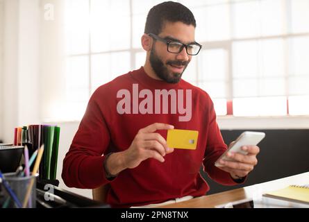 Smiling handsome hispanic man using bank card and phone Stock Photo