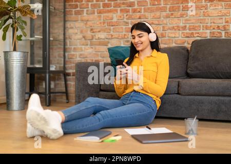 Cheerful teen mexican lady using cellphone wearing headphones texting in social media app, sitting on floor near sofa Stock Photo