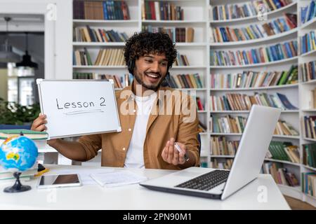 Latin American teacher teaches online students, the man sits in the library of the academic university with books, shows the subject of the lesson to the laptop camera. Stock Photo
