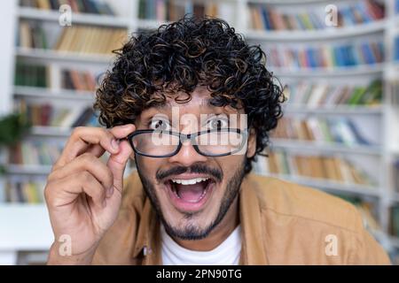 Surprised man looking at camera, hispanic close-up smiling and taking off glasses, student inside academic university library. Stock Photo