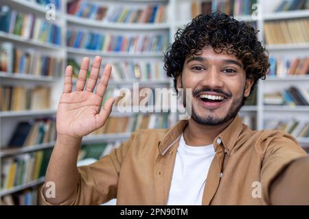 Student video call, hispanic man smiling and looking into phone camera, man using app for online communication with friends, student taking selfie photo in library. Stock Photo