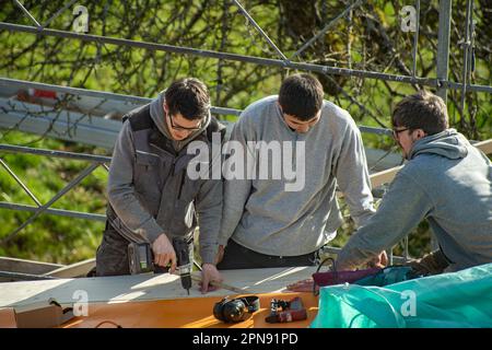 Three young carpenters working on a roof. A carpenter with impact driver screws a screw into a wooden board. Apprentice learns from trainer. Stock Photo