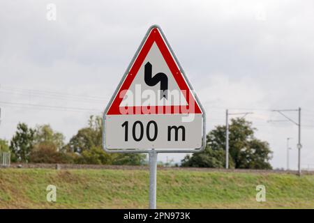 swiss traffic sign: double bend starting to the left. in 100 meters. cloudy during the day without people. drive slowly situation can be dangerous Stock Photo