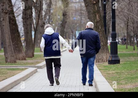 Elderly man and woman walking down the spring street, rear view. Couple with grey hair holding hands together, concept of old age Stock Photo